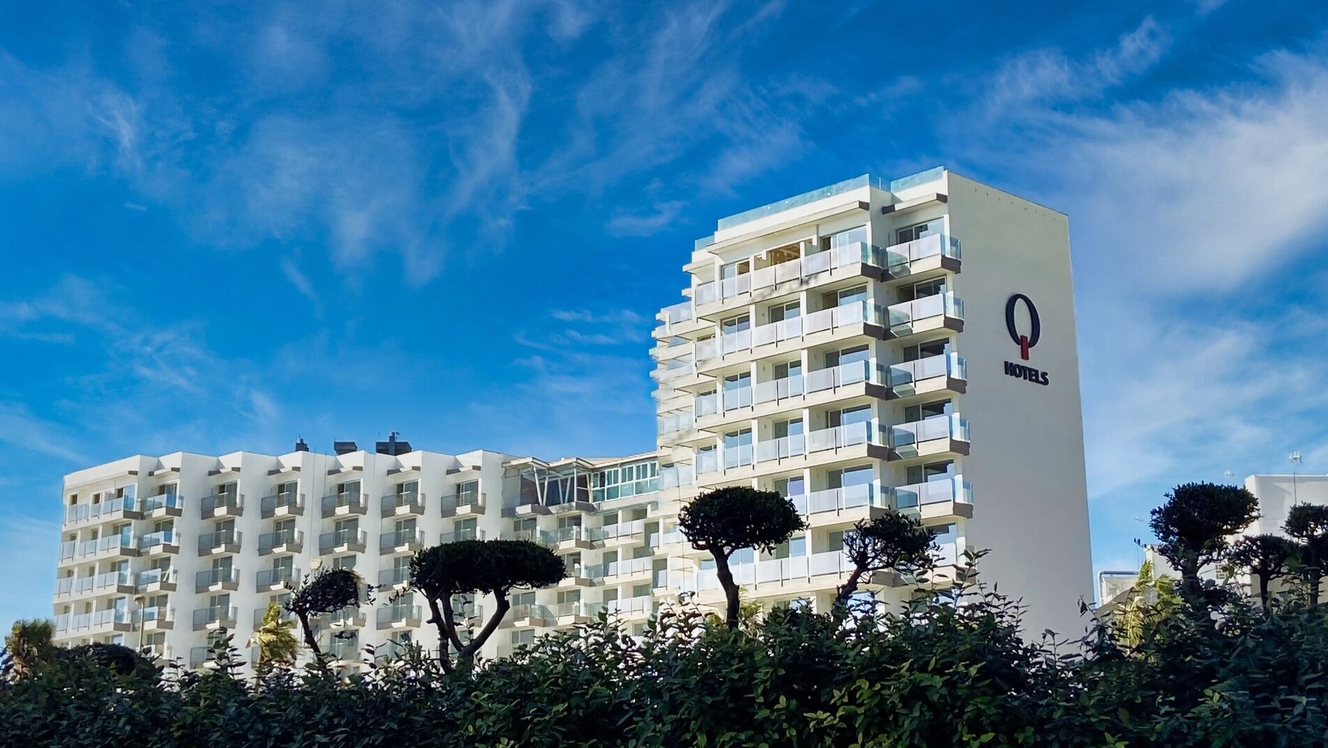 A modern hotel building with multiple floors, balconies, and a prominent logo on the side.