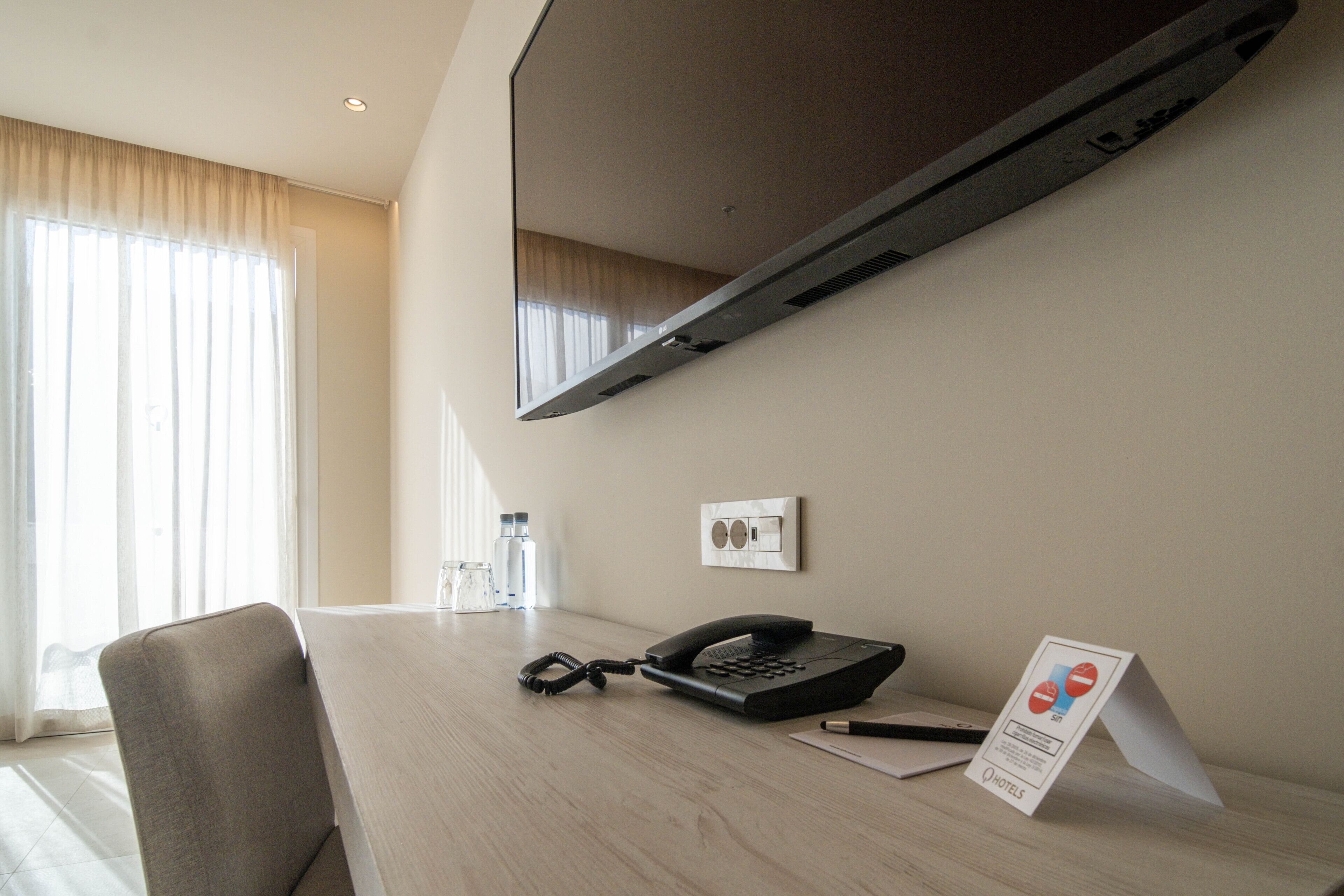 Quarto de hotel com uma escrivaninha, telefone, espelho e janela com cortinas.
