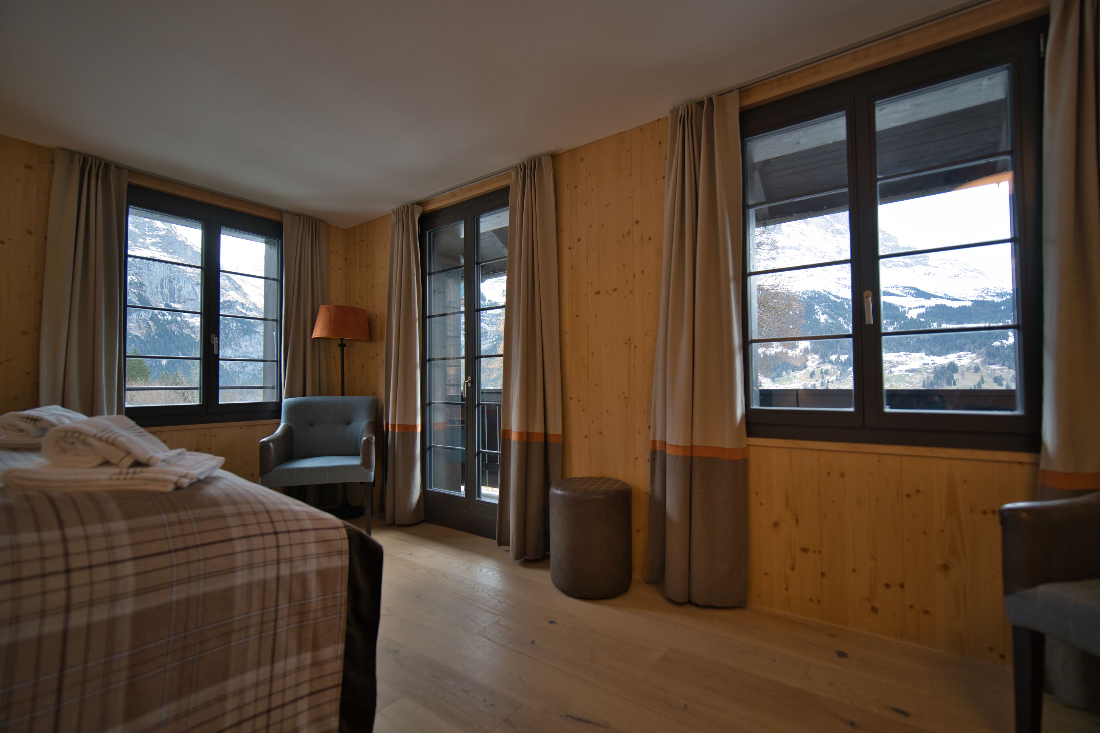 Una habitación con una cama, una silla, una lámpara, una ventana con vistas a montañas y paredes de madera.