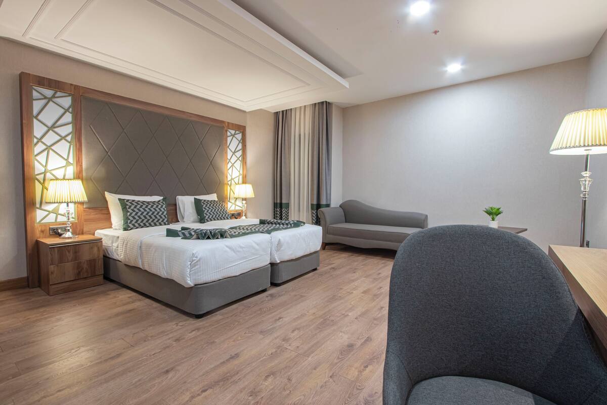 A modern hotel room with a bed, two bedside tables, a sofa, a desk, and a lamp.