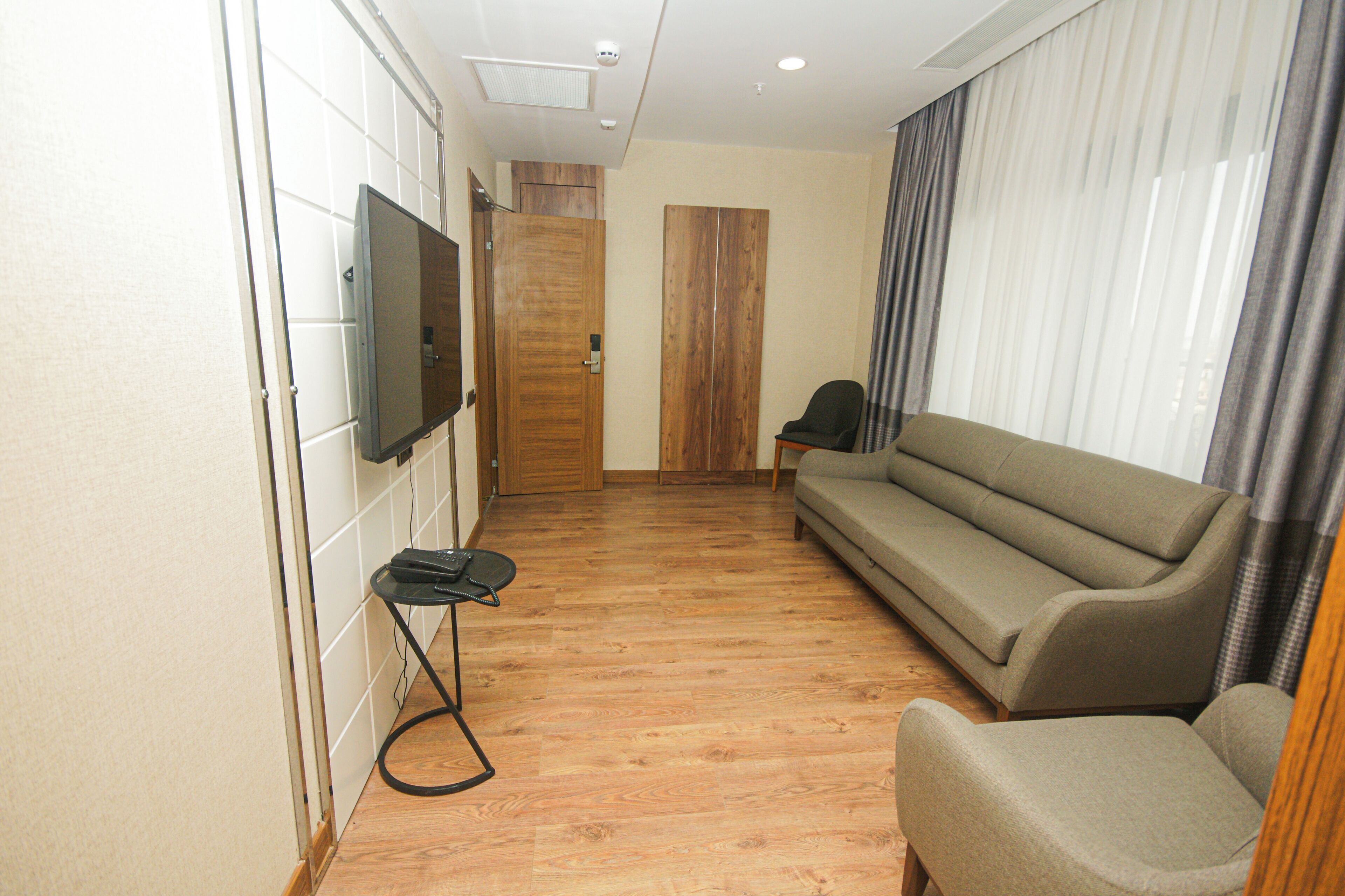 A modern living room with a wooden floor, a grey sofa, a black chair, a TV mounted on the wall, and a wooden door.
