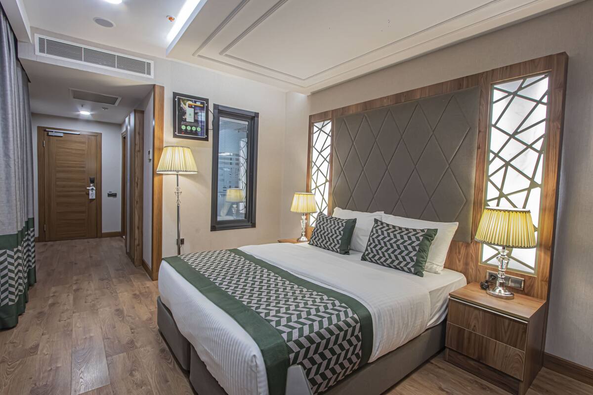 A hotel room with a large bed, two bedside lamps, a headboard with a geometric pattern, a wooden nightstand, and a view through a window with a geometric design.