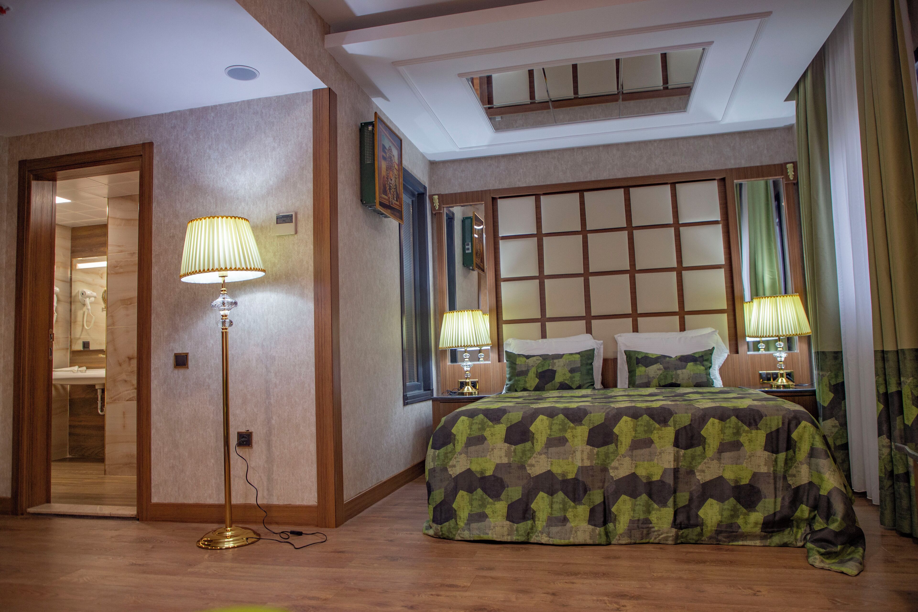 Ein Hotelzimmer mit einem Bett, zwei Nachttischlampen, einer Stehlampe, einem hölzernen Kopfteil und Blick auf ein Badezimmer durch eine geöffnete Tür.