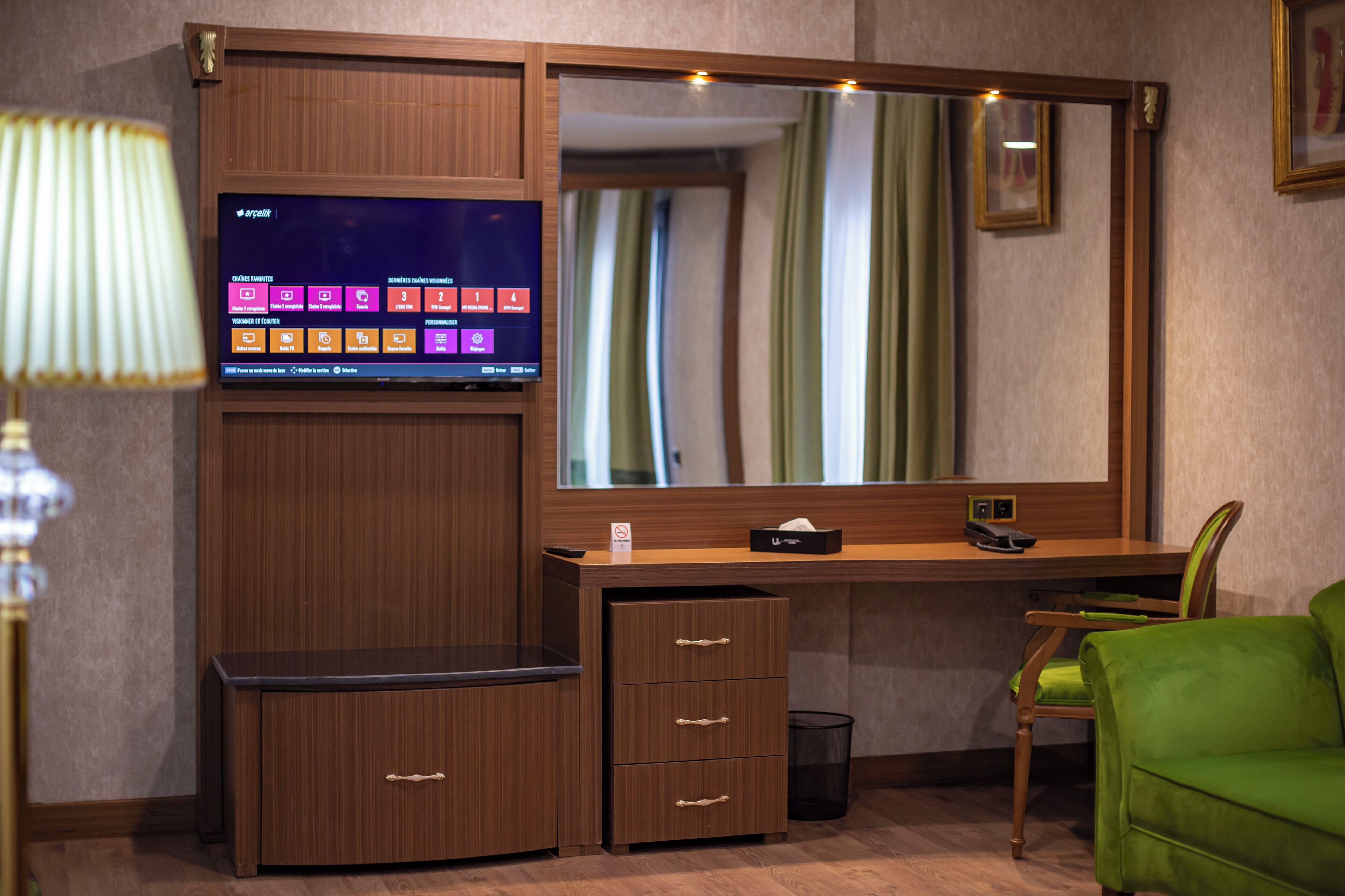 Une chambre d’hôtel avec un bureau en bois, un grand miroir, une chaise verte, une lampe et un téléviseur affichant le menu du service en chambre.