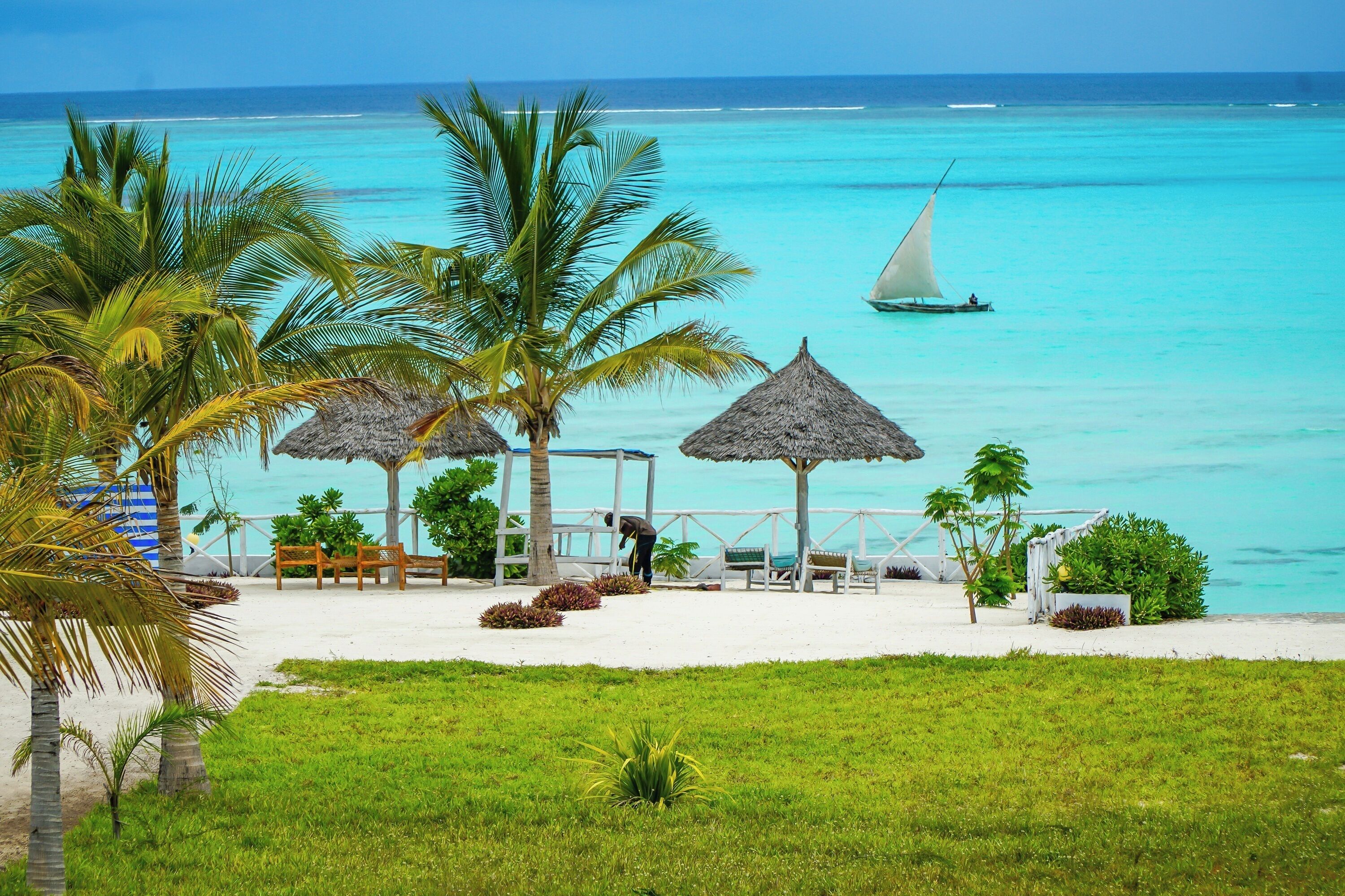 A beach with thatched umbrellas, palm trees, and a sailboat on turquoise waters.