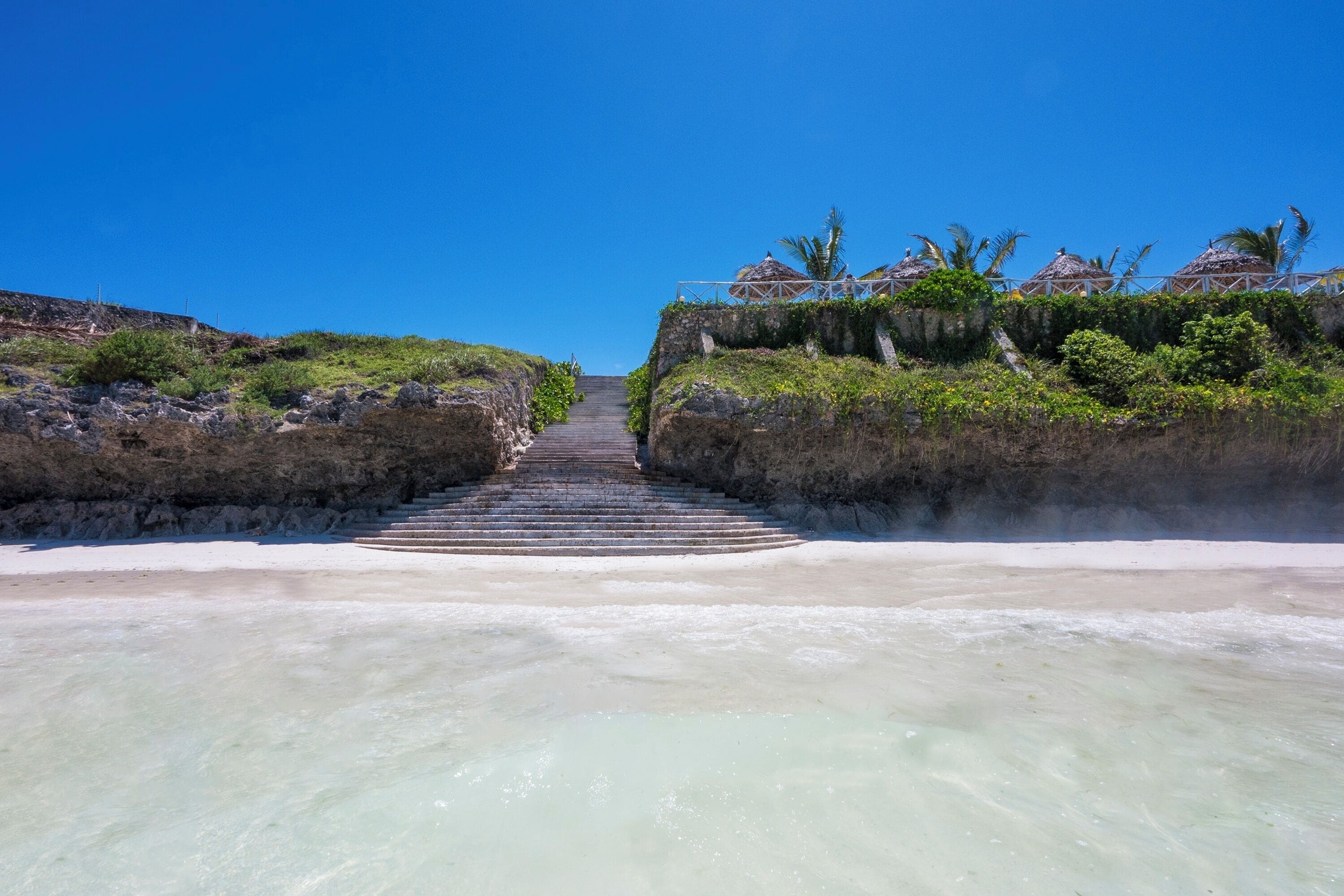 Ein Weg, der zu einem Strand mit klarem Wasser, Stufen und Palmen führt.