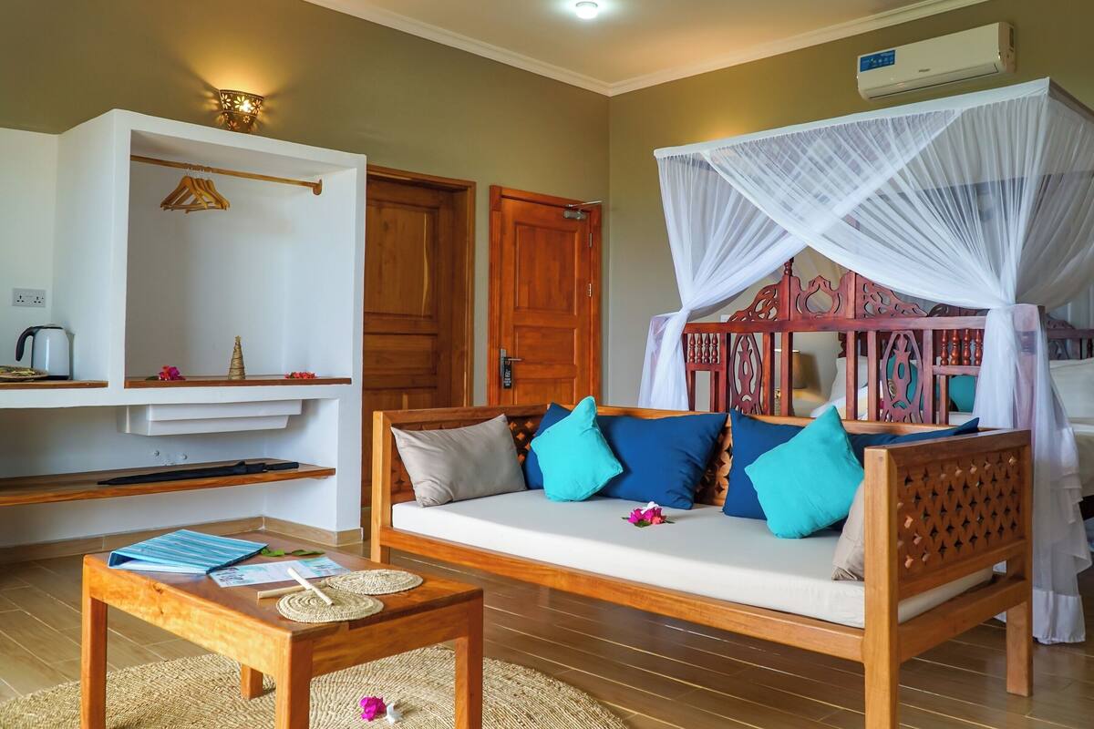 A room with a wooden sofa bed under a canopy, a wooden table with a placemat, a shelf with hangers, and a wall-mounted air conditioner.