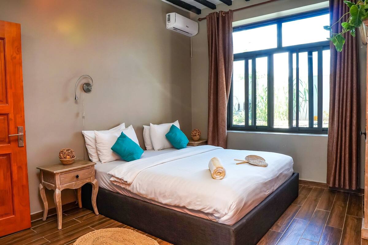 A bedroom with a large bed, a wooden nightstand, a wall-mounted air conditioner, and a window with curtains.