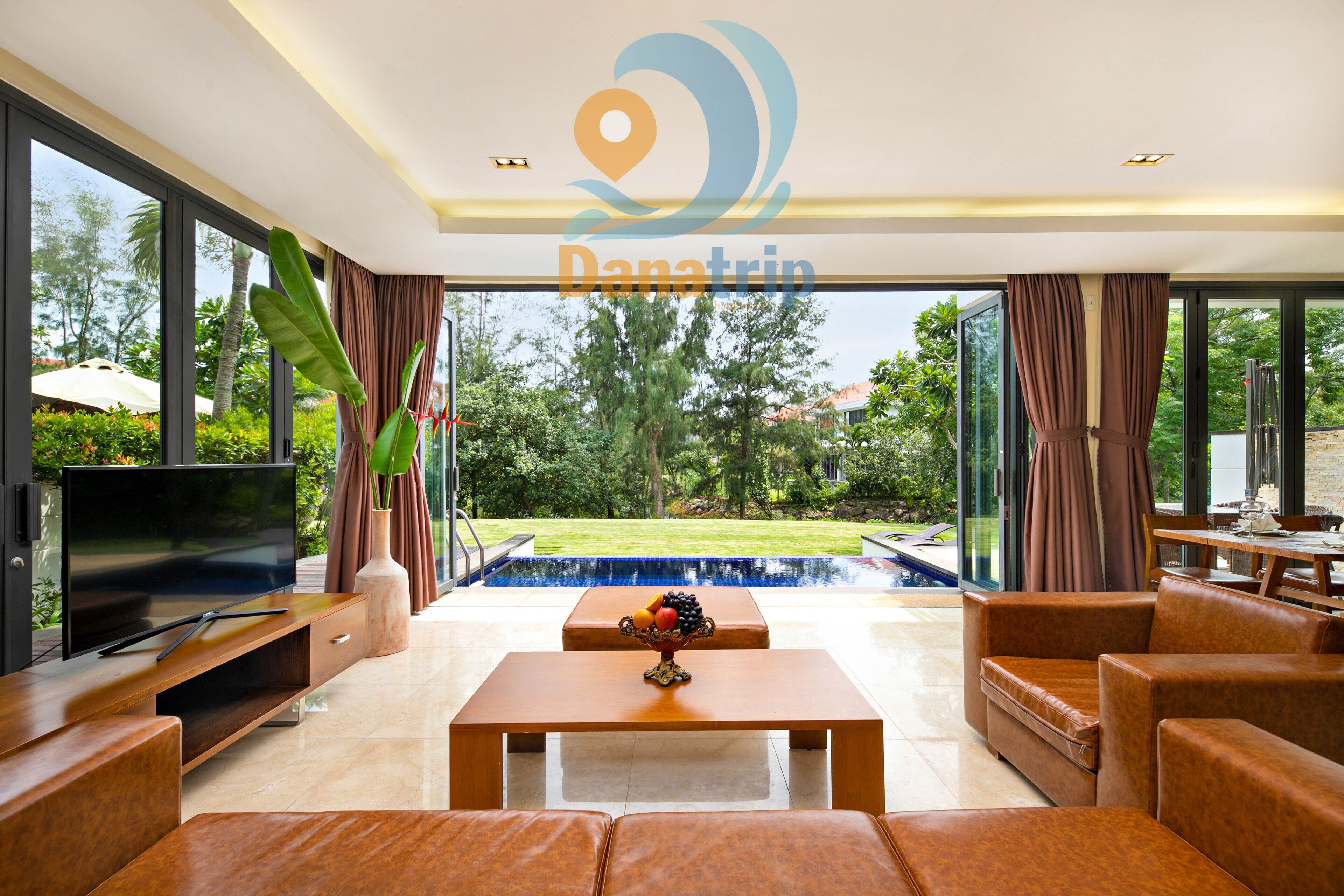 A modern living room with a TV, comfortable seating, a coffee table, and a view of a swimming pool and greenery.