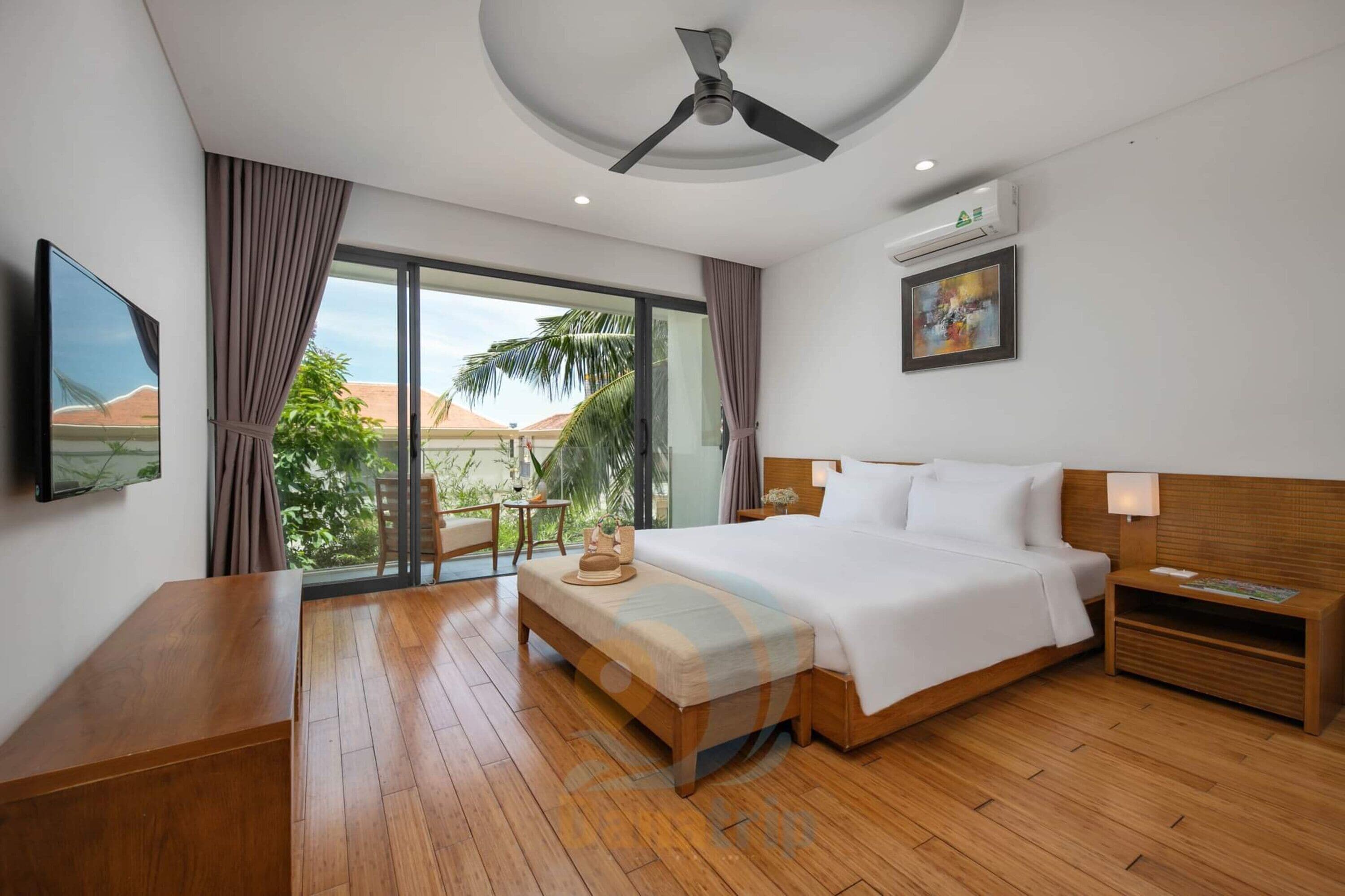 Habitación de hotel con una cama grande, piso de madera, ventilador de techo, televisión, ventana con cortinas y vistas a palmeras y montañas.
