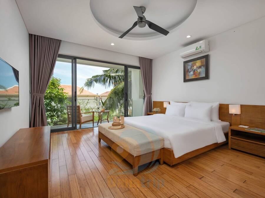 Habitación de hotel con una cama grande, piso de madera, ventilador de techo, televisión, ventana con cortinas y vistas a palmeras y montañas.
