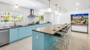 A modern kitchen with light blue cabinets, a central island with bar stools, and a painting of a car on the wall. - Blue Door at Indian Canyons – Mid-Century Retreat with Pool, Spa, Golf Course and Mountain Views (Palm Springs)