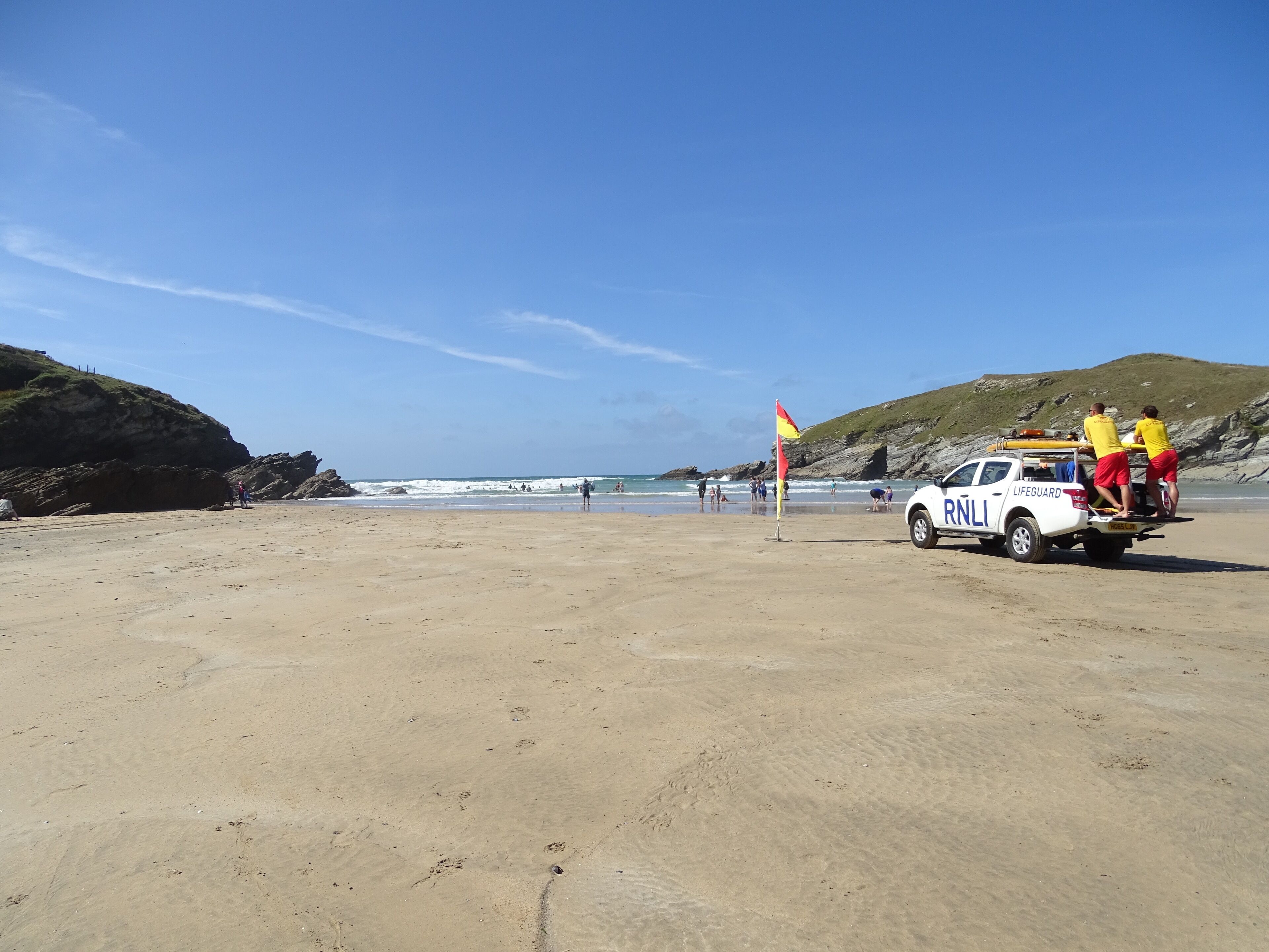 A lifeguard station with a white vehicle on a sandy beach, a flag, and rocky hills in the background.