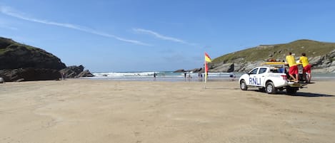 A lifeguard station with a white vehicle on a sandy beach, a flag, and rocky hills in the background.