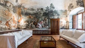 Ein Zimmer mit Wandgemälde, zwei weißen Sofas, einem Glastisch und einem Holzschrank.