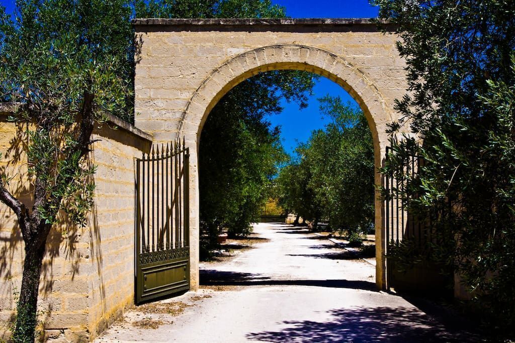 Un arco de piedra con una verja metálica, rodeado de árboles y un cielo despejado.