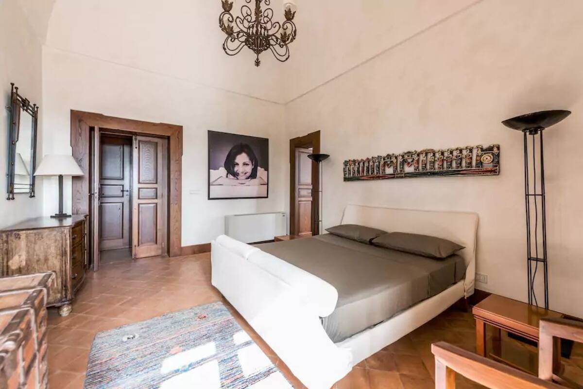A bedroom with a bed, a desk, a chair, a lamp, a chandelier, and a door.