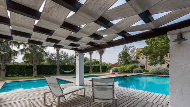Eine Holzterrasse mit zwei weißen Liegestühlen, einem Swimmingpool und einer Pergola darüber.