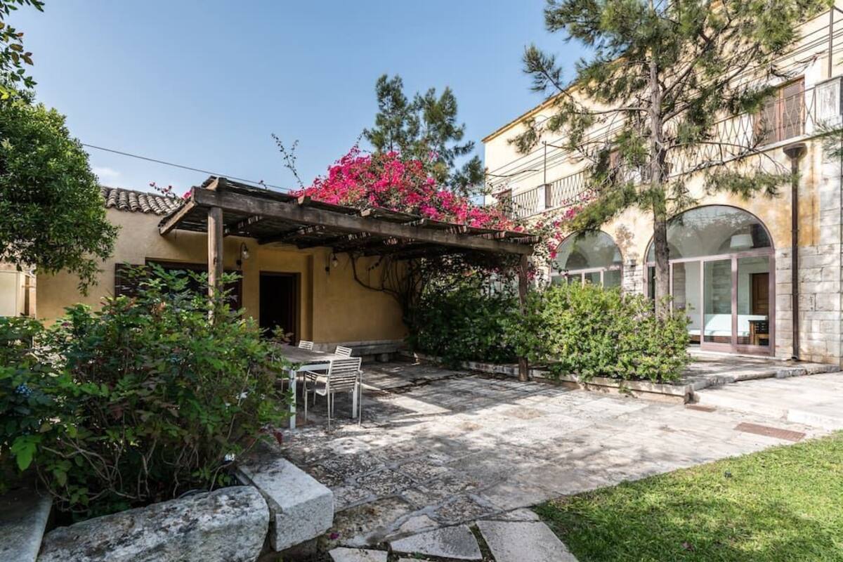 A courtyard with a pergola covered in flowering vines, stone seating, and a stone pathway leading to a building with an arched entrance.
