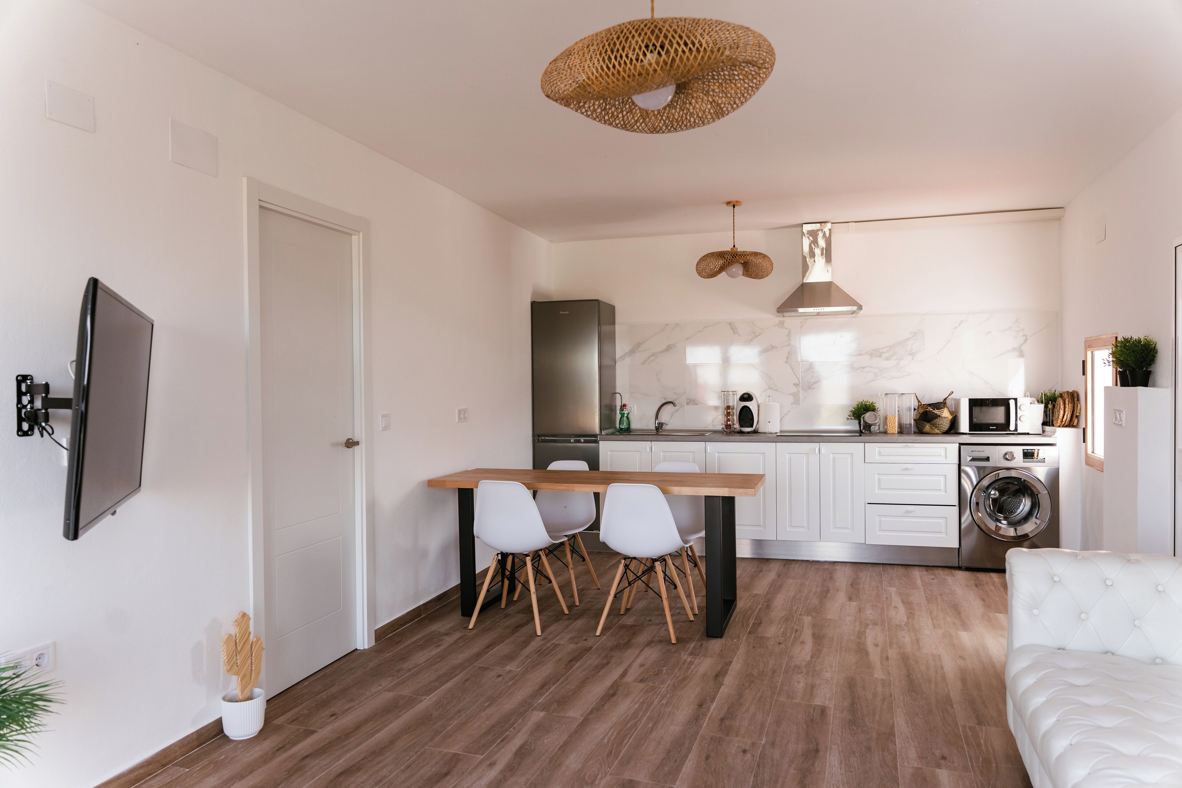 Una cocina moderna con armarios blancos, un refrigerador de acero inoxidable, un horno integrado y una lavadora. Hay una mesa de comedor con sillas y una lámpara colgante.