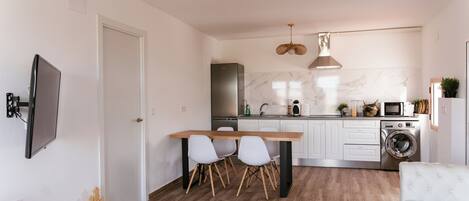 Una cocina moderna con armarios blancos, un refrigerador de acero inoxidable, un horno integrado y una lavadora. Hay una mesa de comedor con sillas y una lámpara colgante.