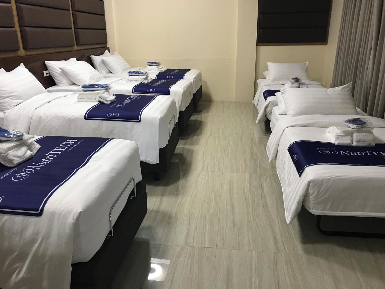 A hotel room with three beds, each with a white bedspread and a blue headboard. The room has a wooden floor and a headboard with a padded design.
