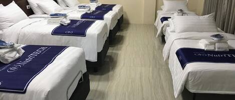 A hotel room with three beds, each with a white bedspread and a blue headboard. The room has a wooden floor and a headboard with a padded design.