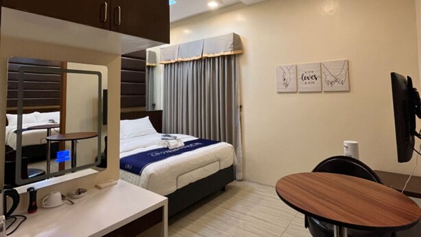 A hotel room with a bed, a desk, a chair, a mirror, a TV, and artwork on the wall. - NutriTECH Hotel and Events (Calapan)