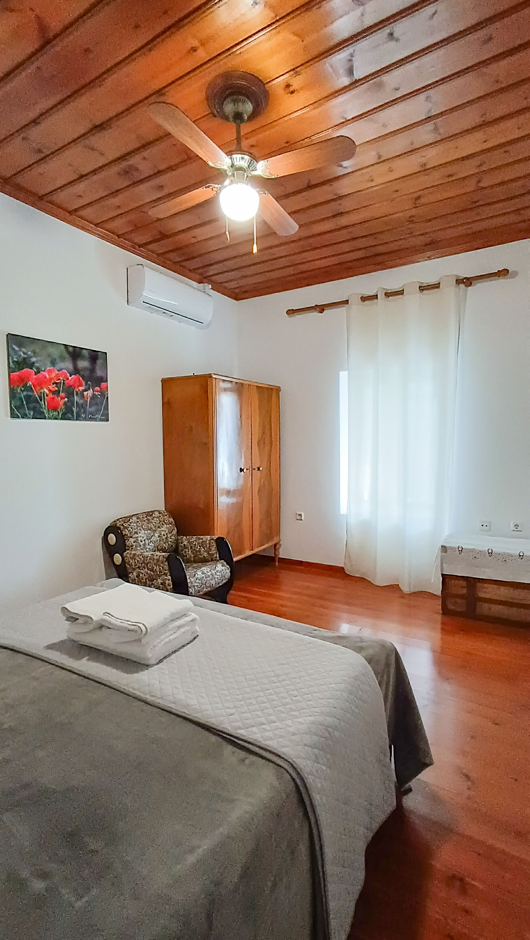 A room with a bed, a wooden wardrobe, a ceiling fan, and a painting on the wall.