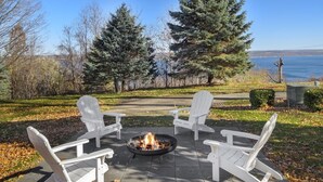 A patio with four Adirondack chairs arranged around a fire pit, overlooking a body of water and trees.
