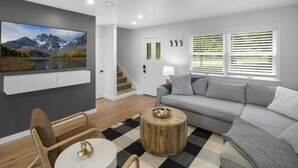 A modern living room with a grey sofa, wooden chairs, a round coffee table, and a large painting of a mountain landscape.