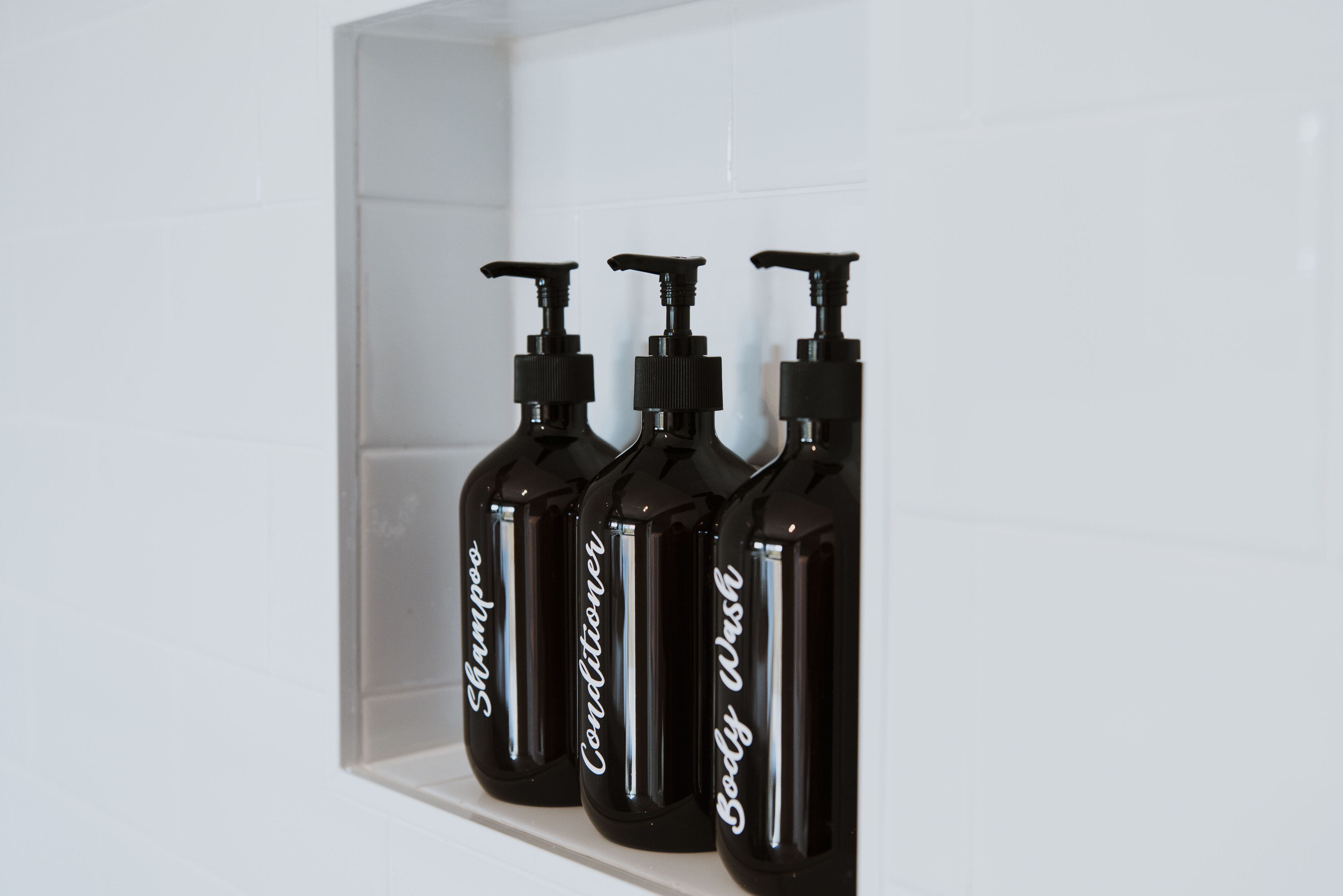 Three black bottles labeled shampoo, conditioner, and body wash on a white shelf.