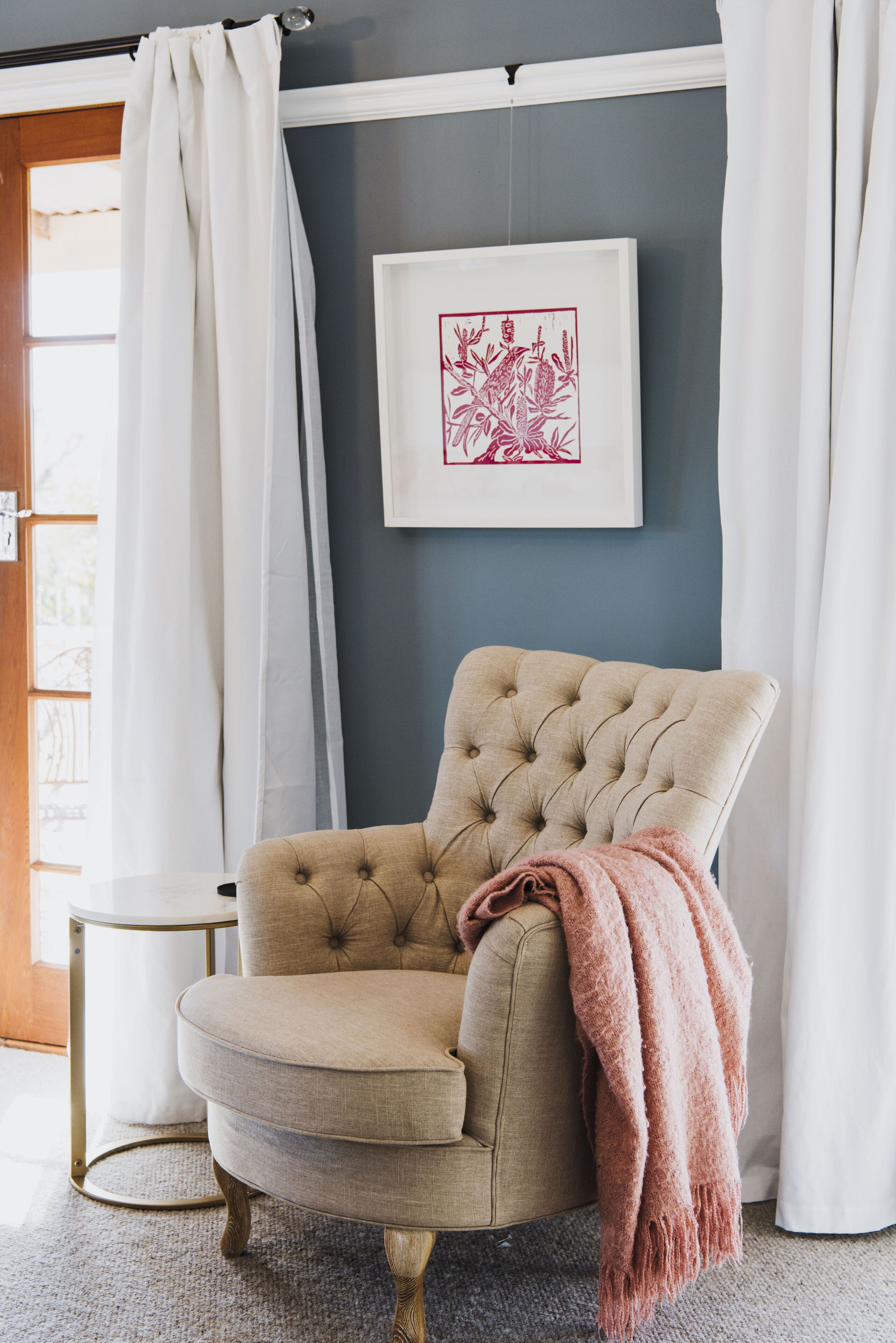A tufted armchair with a pink throw blanket, a framed artwork on the wall, and a window with curtains.