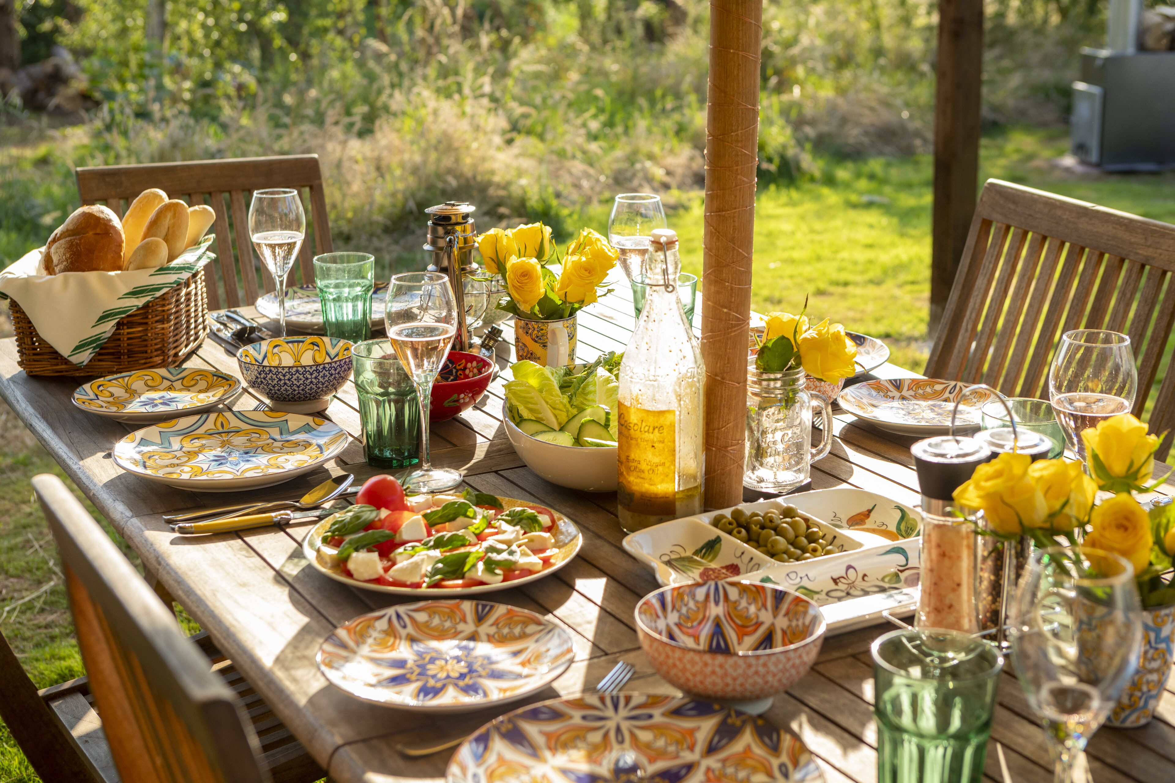 A wooden table set outdoors with various dishes, glasses, and a bottle of wine.