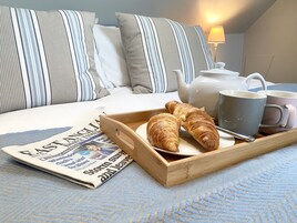 Una cama bien hecha con almohadas a rayas, una bandeja con cruasanes, una tetera y dos tazas, junto con un periódico.