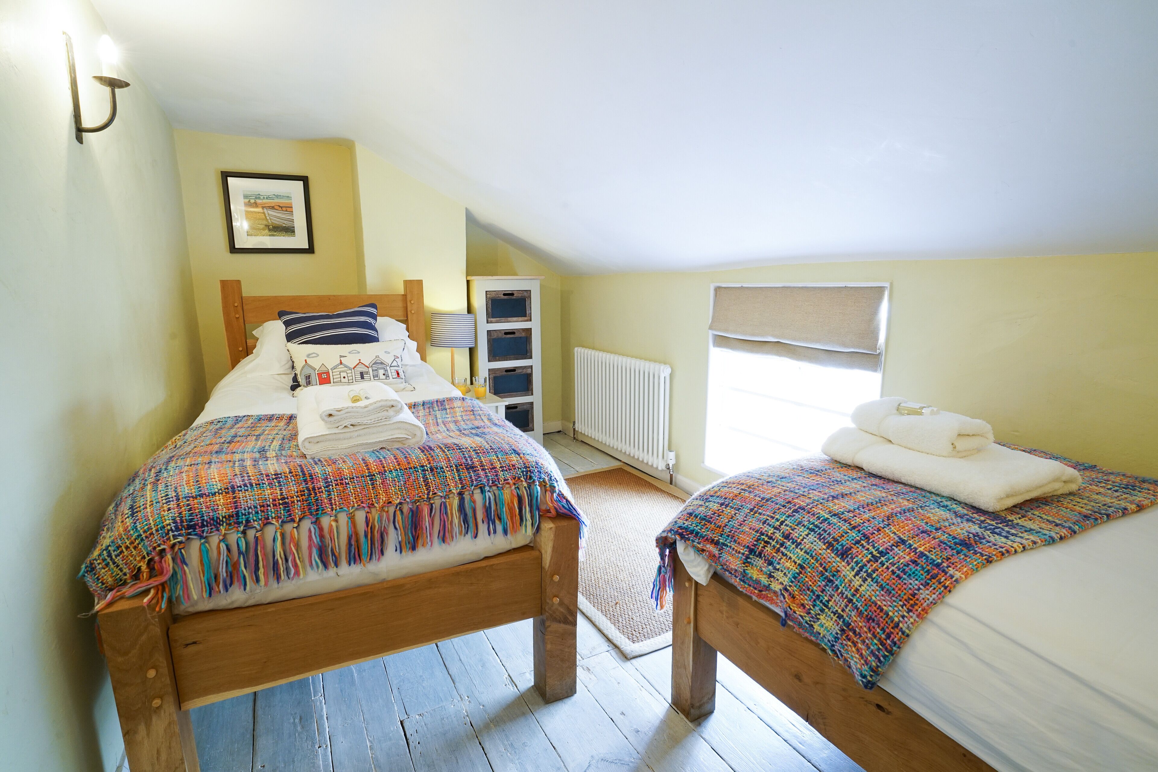 A bedroom with two beds, a radiator, a window with blinds, and a framed picture on the wall.