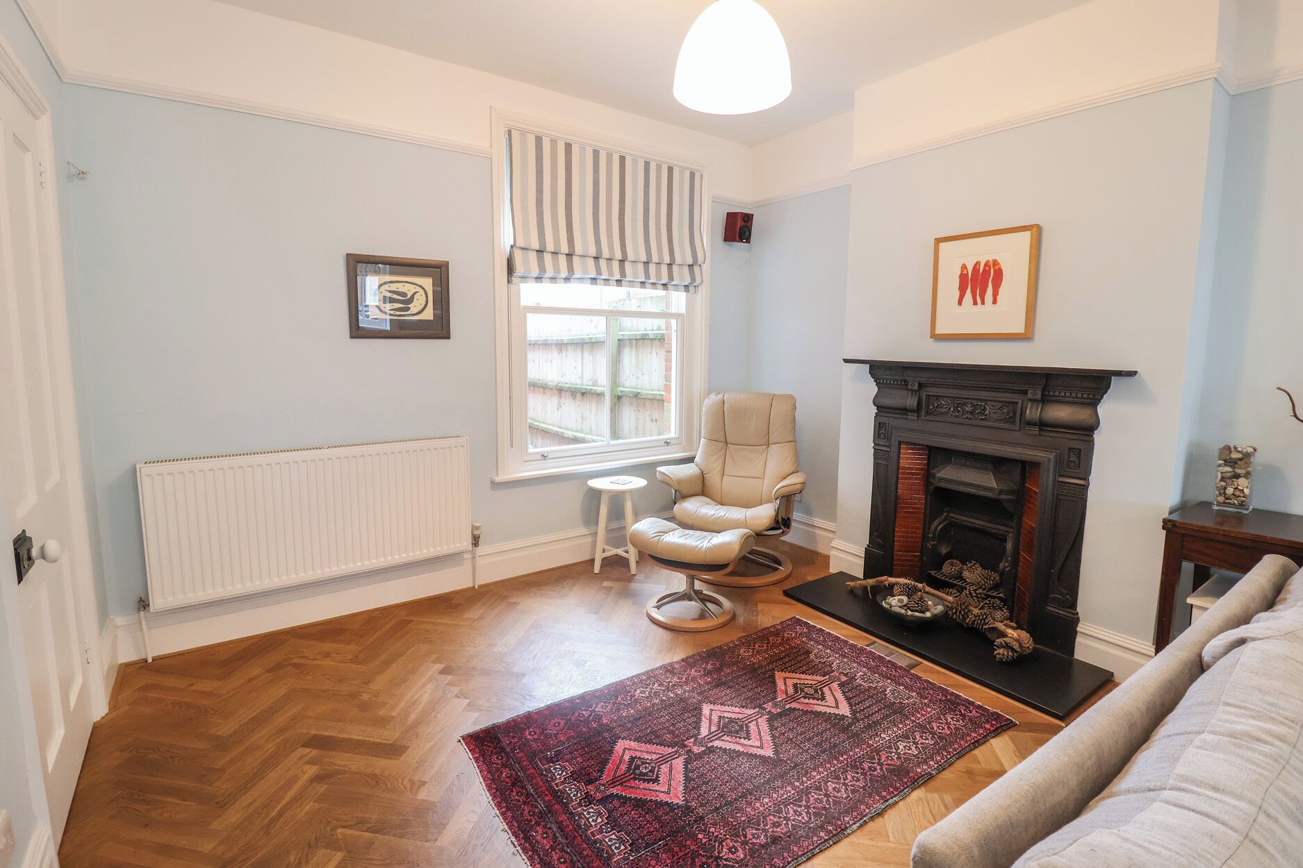 A living room with a fireplace, a radiator, a window with striped curtains, a chair, a sofa, and a patterned rug.