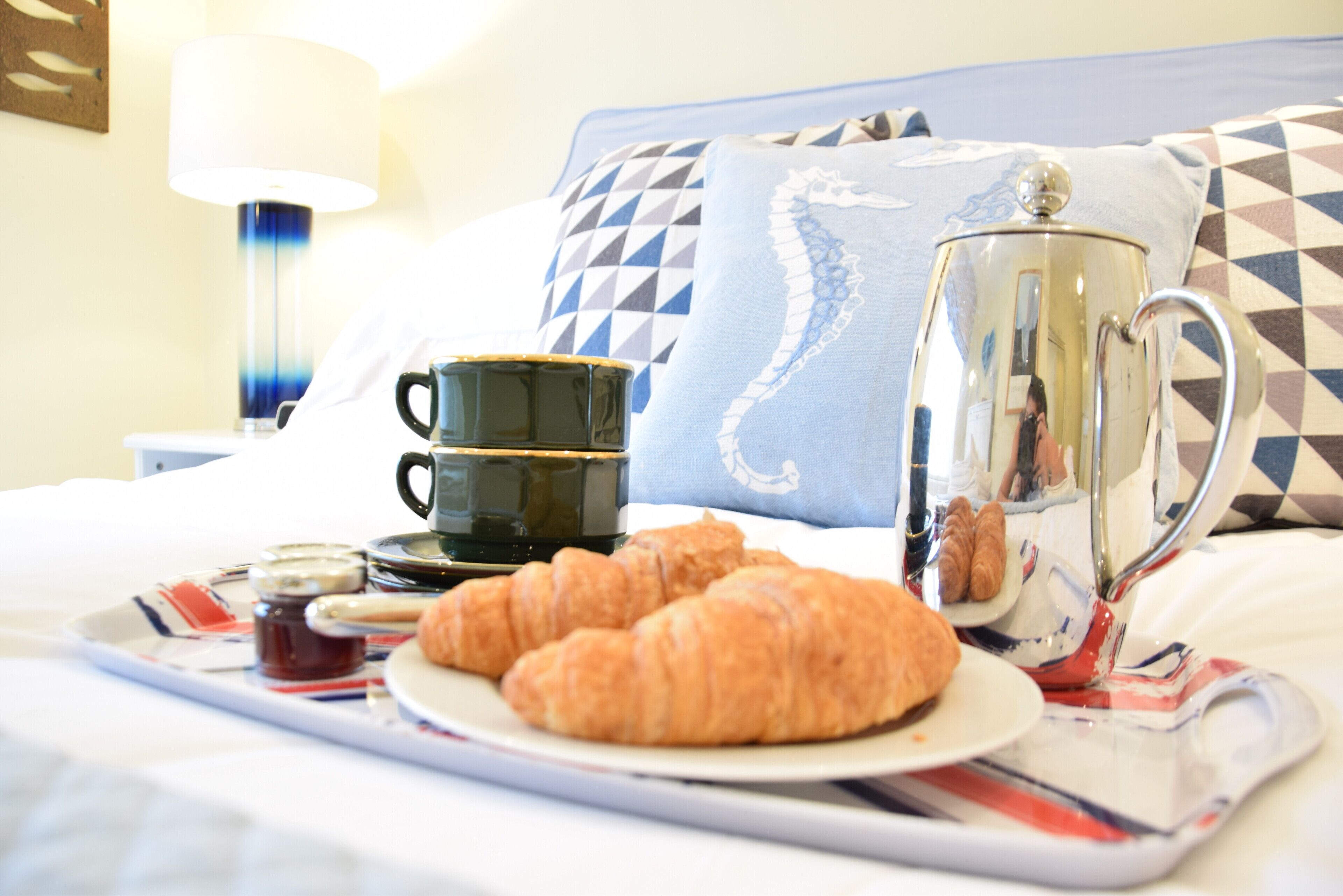 Un plateau de petit-déjeuner avec des croissants, une cafetière et des tasses, posé sur un lit agrémenté d’un coussin à motifs et d’une lampe en arrière-plan.