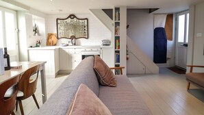 A modern kitchen with white cabinetry, a dining area with a wooden table and chairs, a grey sofa with cushions, and a staircase leading to another level. - Mariners Cottage (Aldeburgh)