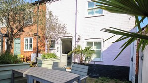 An outdoor patio with a table and chairs, a small garden area with plants, and a brick building with a white door. - Mariners Cottage (Aldeburgh)
