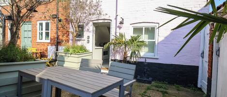 An outdoor patio with a table and chairs, a small garden area with plants, and a brick building with a white door.