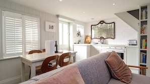 A modern kitchen with white cabinetry, a dining area with a table and chairs, and a living room with a sofa and a bookshelf. - Mariners Cottage (Aldeburgh)