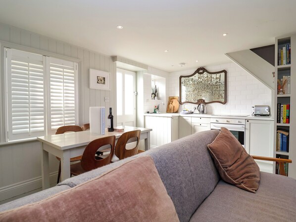 A modern kitchen with white cabinetry, a dining area with a table and chairs, and a living room with a sofa and a bookshelf. - Mariners Cottage (Aldeburgh)