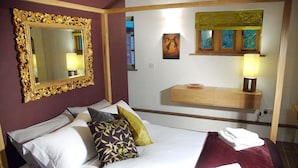 Habitación de hotel con un espejo grande y decorativo, una cama con sábanas blancas y almohadas estampadas, una mesita de noche de madera con una lámpara y una ventana pequeña con vistas a la vegetación.
