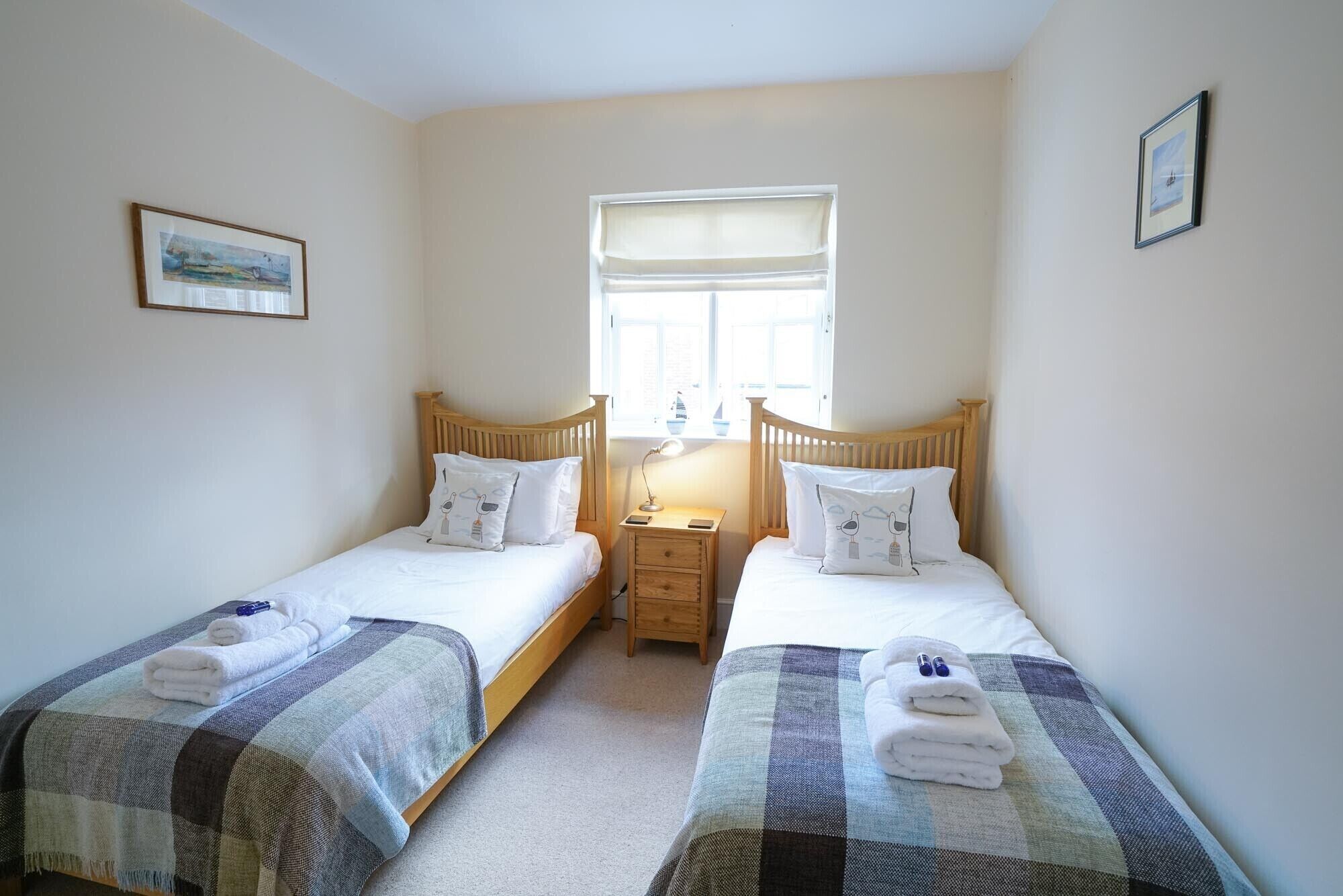 Une chambre avec deux lits simples, chacun doté d’une tête de lit en bois, d’une petite table de chevet et d’une lampe. Il y a une fenêtre avec un store blanc, et les murs sont peints dans une couleur claire.