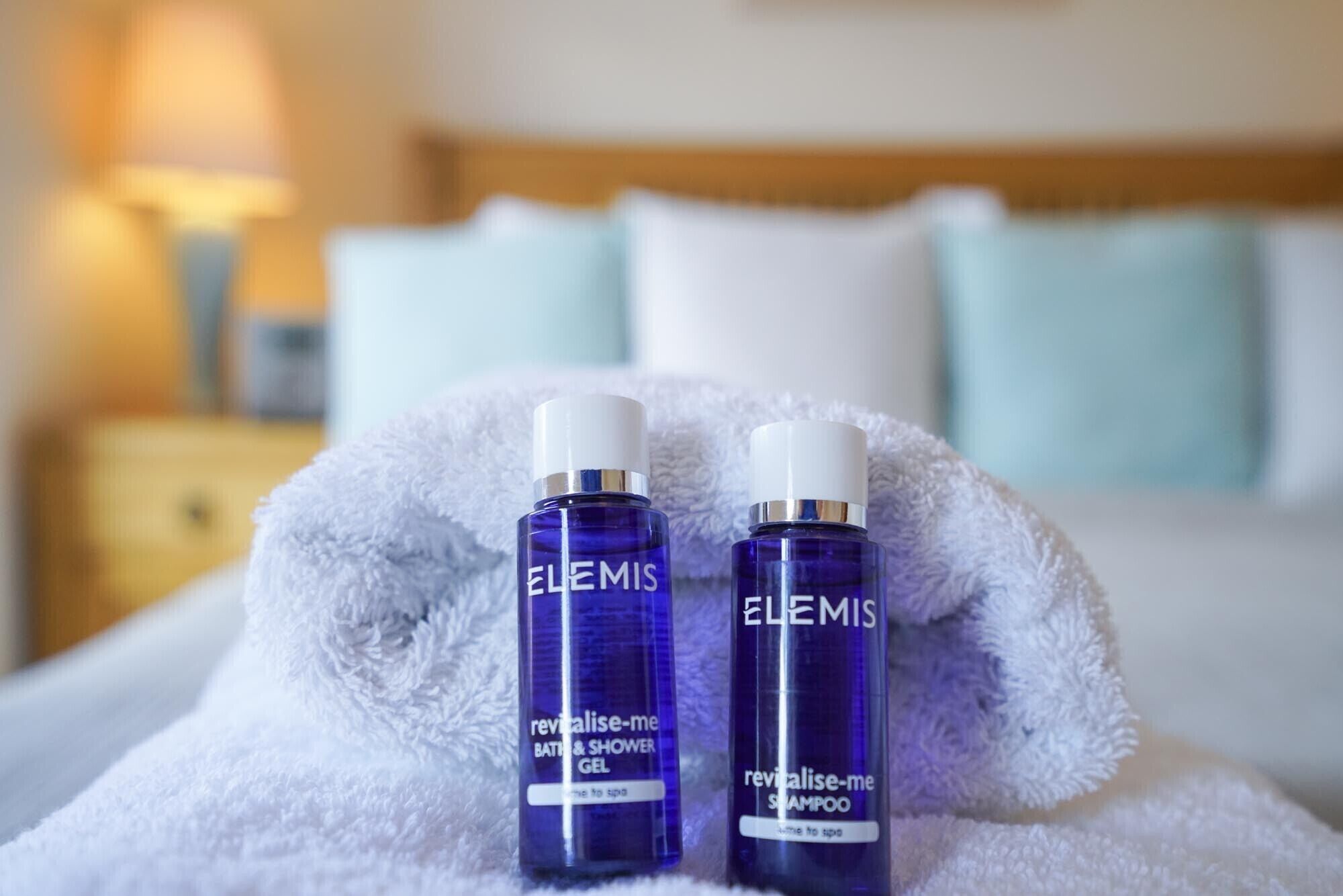 Duas embalagens de Elemis Revitalise-me Gel de Banho e Shampoo sobre uma toalha branca numa cama.