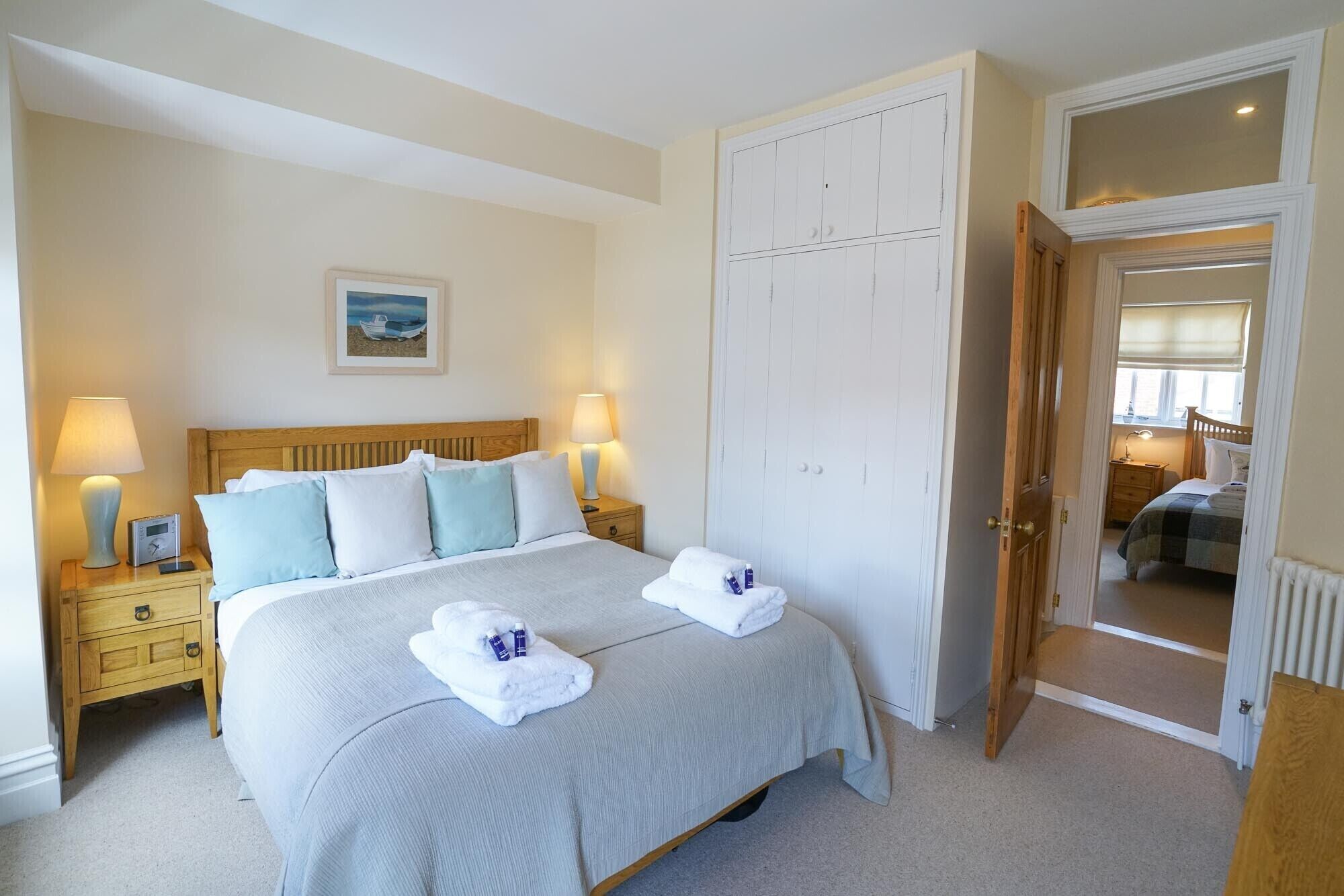 A neatly arranged bedroom with a bed, bedside lamps, a nightstand, a wooden headboard, a white wardrobe, and a door leading to another room.