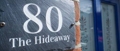 A slate house number sign with "80 The Hideaway" on a brick wall.