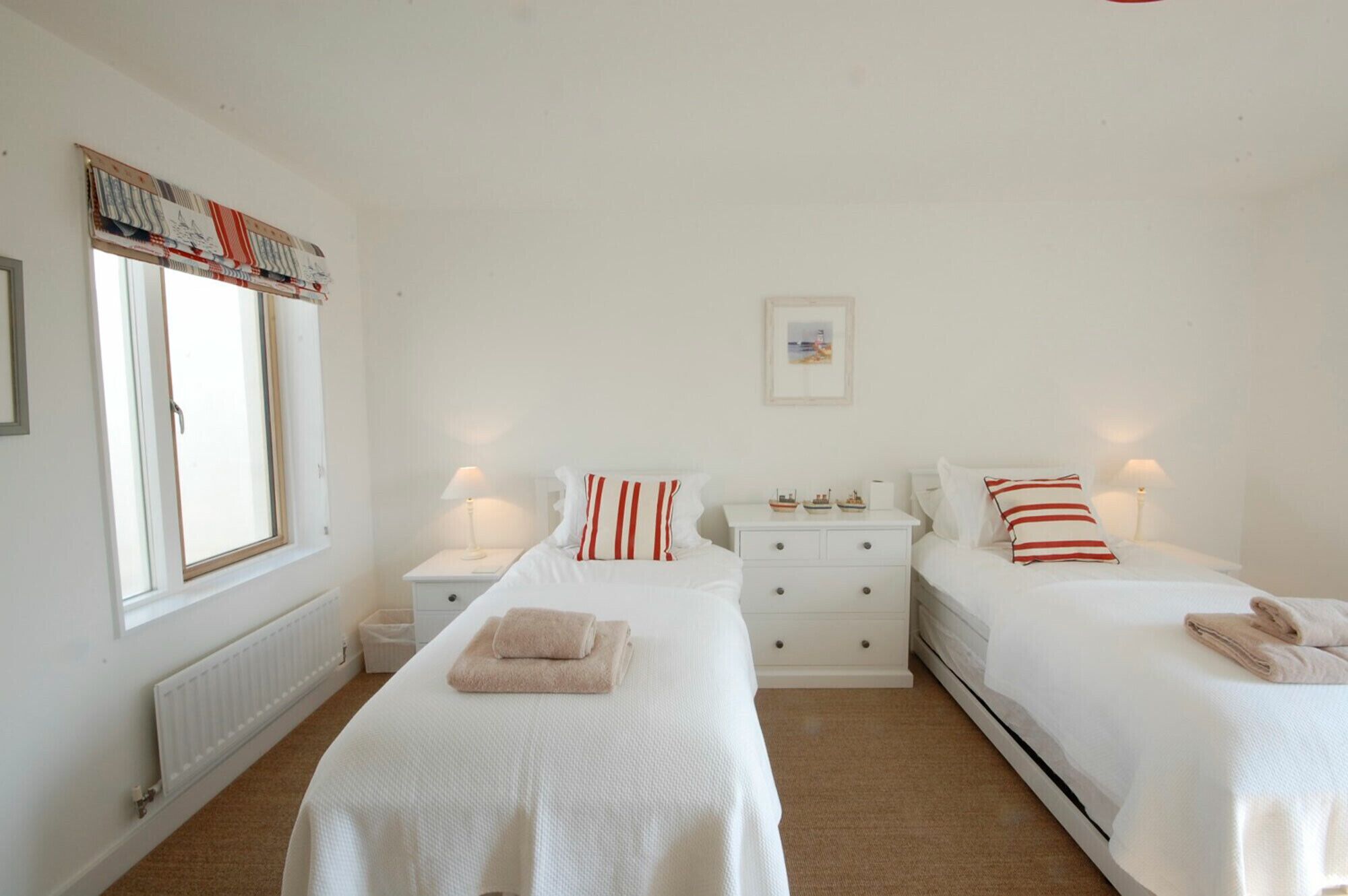 Un dormitorio con dos camas, ropa de cama blanca, una ventana con cortinas, un radiador y una mesita de noche pequeña con una lámpara.