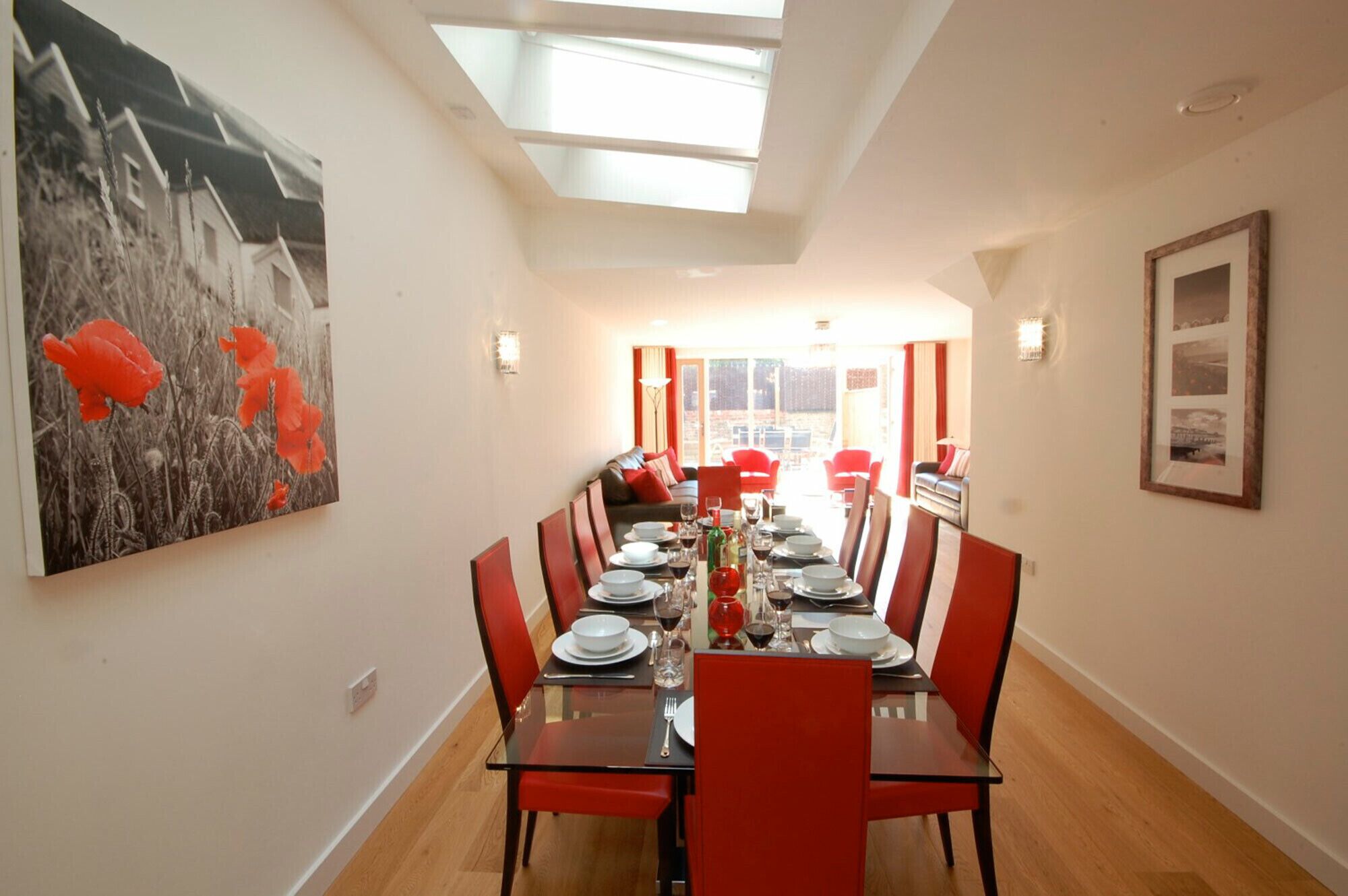 Un comedor con una mesa larga dispuesta para seis personas, sillas rojas y un cuadro de flores rojas en la pared.